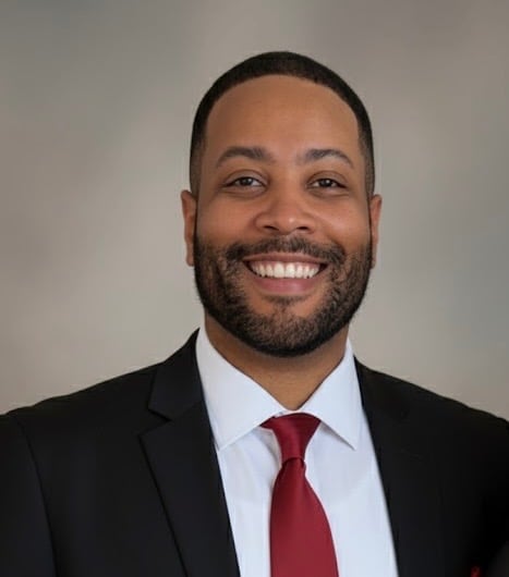 Professional headshot of a smiling man wearing a black blazer, white dress shirt, and burgundy tie against a neutral background.