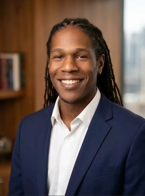 Professional headshot of a man with long dreadlocks wearing a navy blazer and white shirt, smiling at the camera in an office setting.