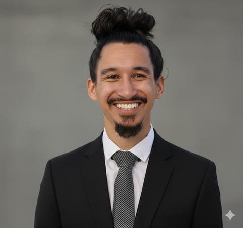 Smiling man with black hair in a top bun, goatee, wearing a black blazer, white dress shirt, and gray tie against a neutral gray background.