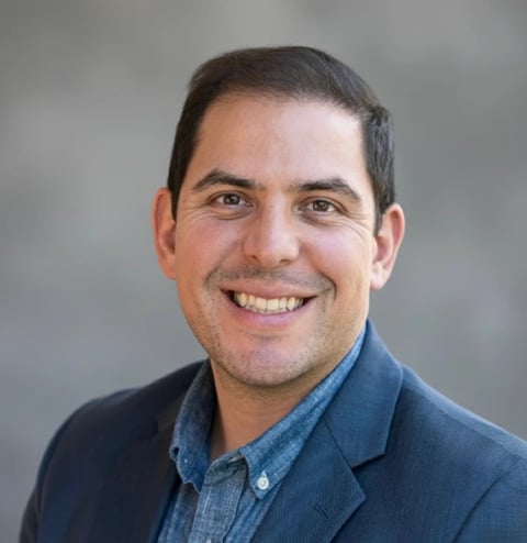 Smiling man with dark hair wearing a blue denim button-up shirt against a gray background