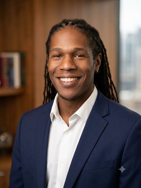 Professional portrait of a smiling man with dreadlocks wearing a navy blazer and white shirt in an office setting