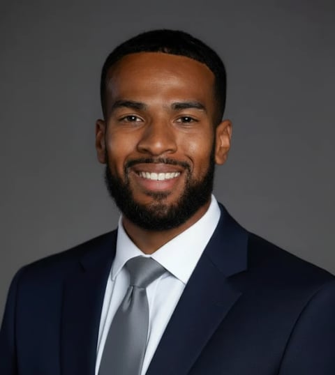 Professional headshot of a man with a beard wearing a dark navy blazer, light blue shirt, and gray tie against a gray background