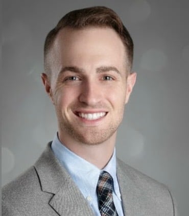 Professional headshot of a smiling man wearing a gray blazer, light blue shirt, and plaid tie against a gray background